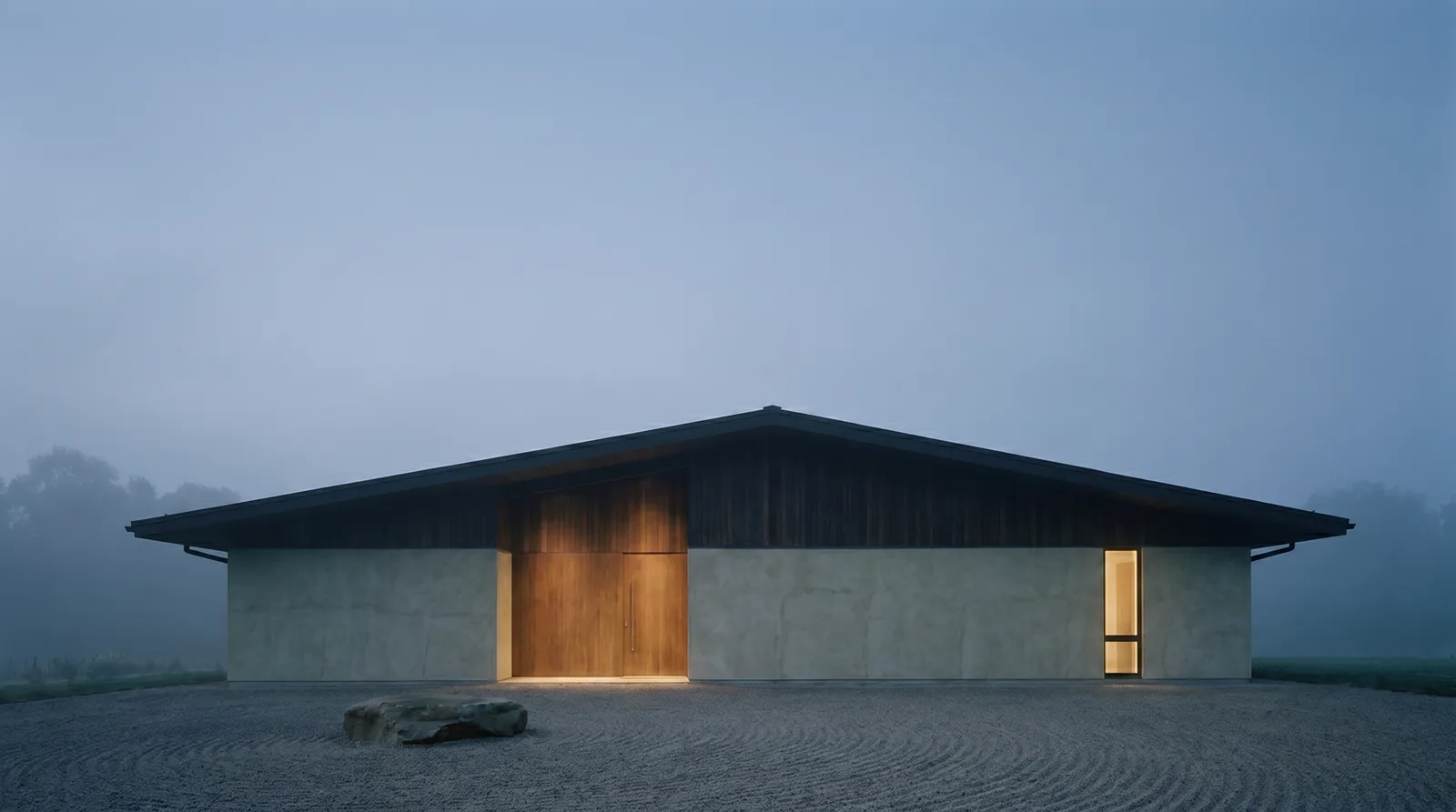The same low house at dusk in thin mist, its plaster walls blue-grey, warm light showing through the timber entrance and one narrow window.