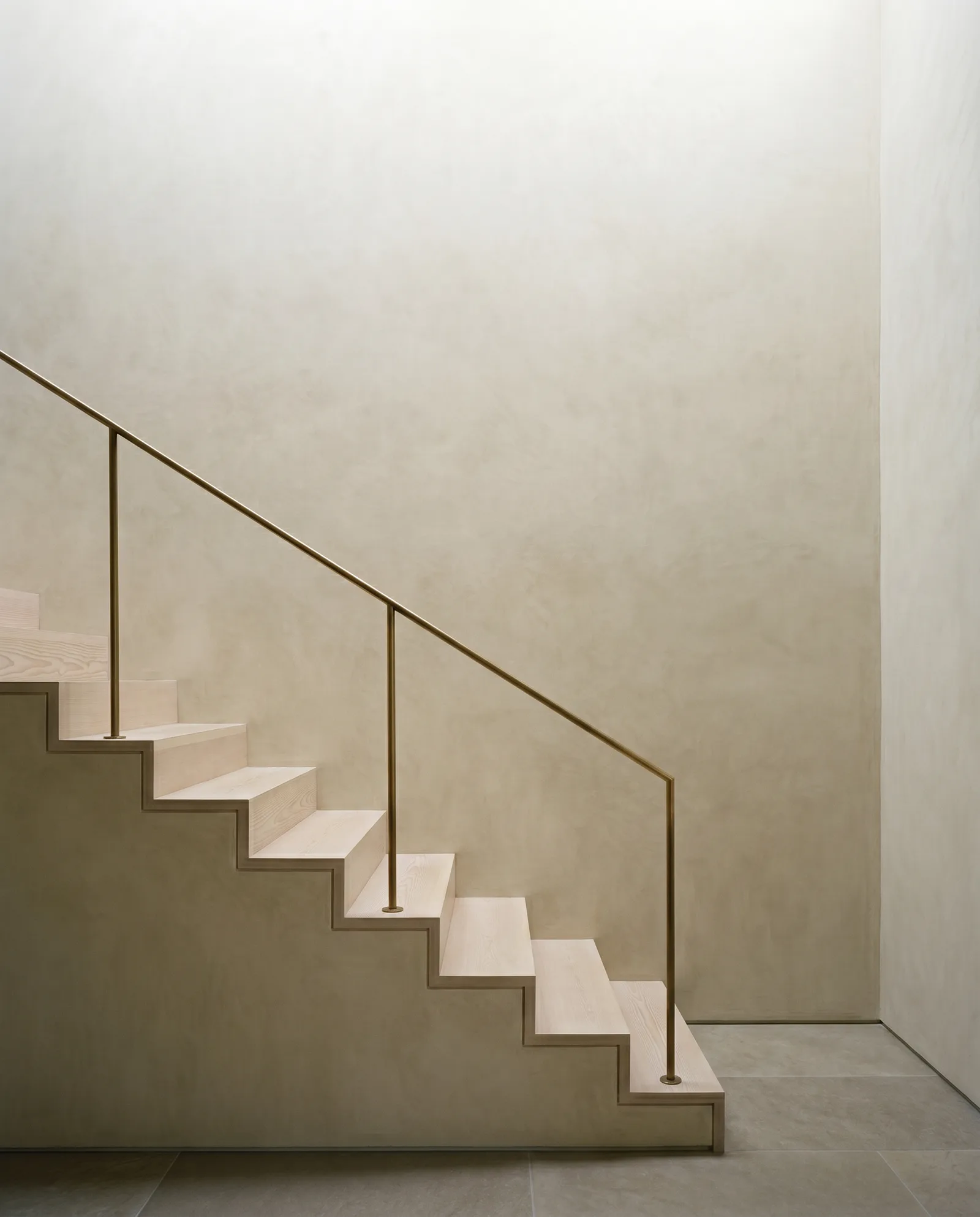 A pale fir stair rising along a lime plaster wall, held by a thin bronze handrail on slender posts.